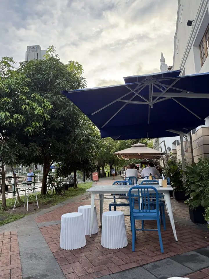 A small dog sitting calmly beside its owner at the outdoor pavement seating of Good Boy George on Rodyk Street in Robertson Quay, Singapore, with the bistro's warm interior visible through the open shopfront.