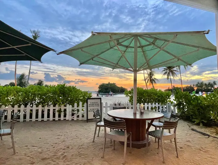 An outdoor dining table under a large parasol overlooking a sandy beach at sunset.